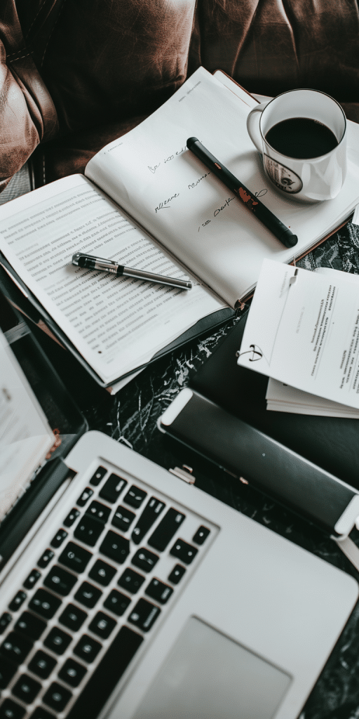 cozy workspace featuring a laptop, open books with handwritten notes, pens, and a cup of coffee.
